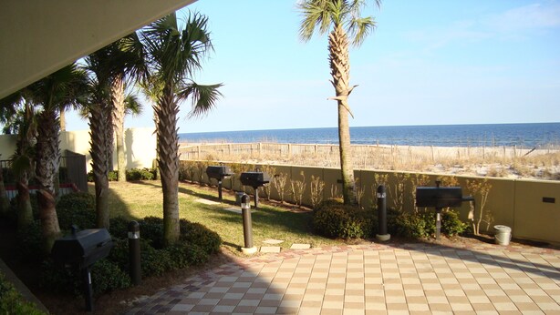 A patio with a tiled floor, a round table, and two chairs. There are palm trees and a view of the beach. - Sunny paradise! Beachfront, bay views & all the amenities! Welcome families! (Orange Beach)