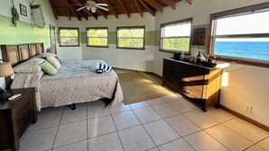 Quarto com cama, escrivaninha, ventoinha no teto e janelas com vista para o oceano.