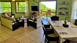 Sala de estar com sofá, mesa de jantar e cadeiras. Há uma televisão na parede e um ventilador de teto.