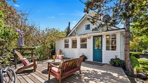 A wooden deck with a bench and chairs, a small table with a vase of flowers, and a house with a blue door and windows. - Bluebird Day - Jefferson Cottage Cozy two-bedroom, perfect for family getaways. (Bend)