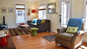 A living room with a sofa, armchair, and a small table. There are windows with blinds, a lamp, and a guitar on the wall. - Bluebird Day - Jefferson Cottage Cozy two-bedroom, perfect for family getaways. (Bend)