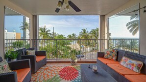 Varanda com móveis de vime, um tapete colorido e vista para palmeiras e edifícios.