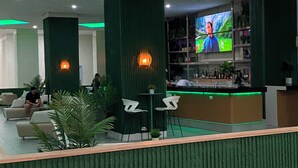 Uma área de recepção com parede verde decorativa, um bar com uma grande tela de TV e assentos com plantas em vasos.
