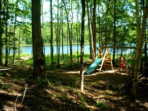 A forested area with a wooden swing set and a lake in the background. - Peaceful Lakefront Lodge w/ Kayaks & No Hidden Fees (Traverse City)