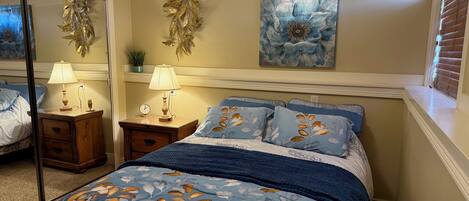 A bedroom with a bed, two bedside tables, a wall-mounted lamp, a clock, a decorative wall piece, and a window with blinds.