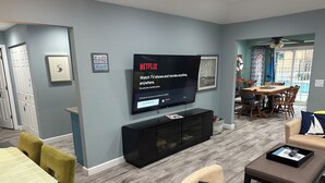 A living room with a flat-screen TV mounted on the wall, a dining area with a wooden table and chairs, and a kitchen area visible through an open door. - Pool and Hot Tub  Beach house 3 bedroom.. 3 bedrooms two bath.   STR19-00084 (Henderson)