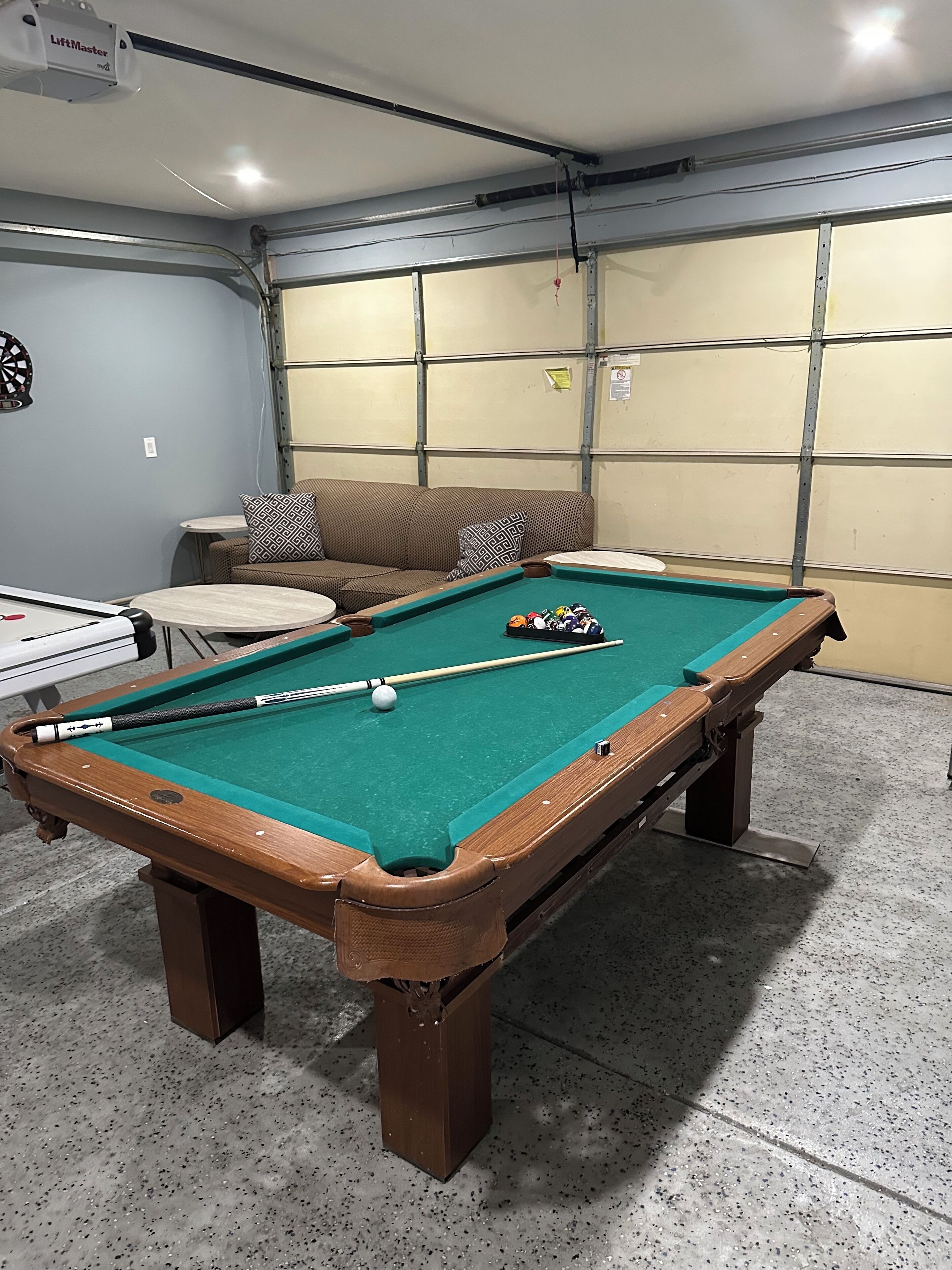 A pool table with a cue stick and balls, a sofa with patterned cushions, and a dartboard.