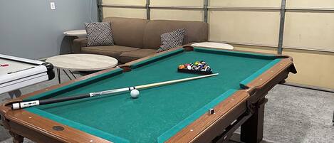 A pool table with a cue stick and balls, a sofa with patterned cushions, and a dartboard.