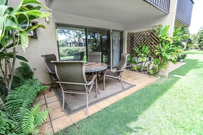 An outdoor patio with a round table and four chairs, surrounded by lush greenery and a view of a residential area. - BEST LOCATION in Kihei! 1BD/1BA by the BEACH! A/C, Clean, Cozy, Wifi, Beach Gear (Kihei)