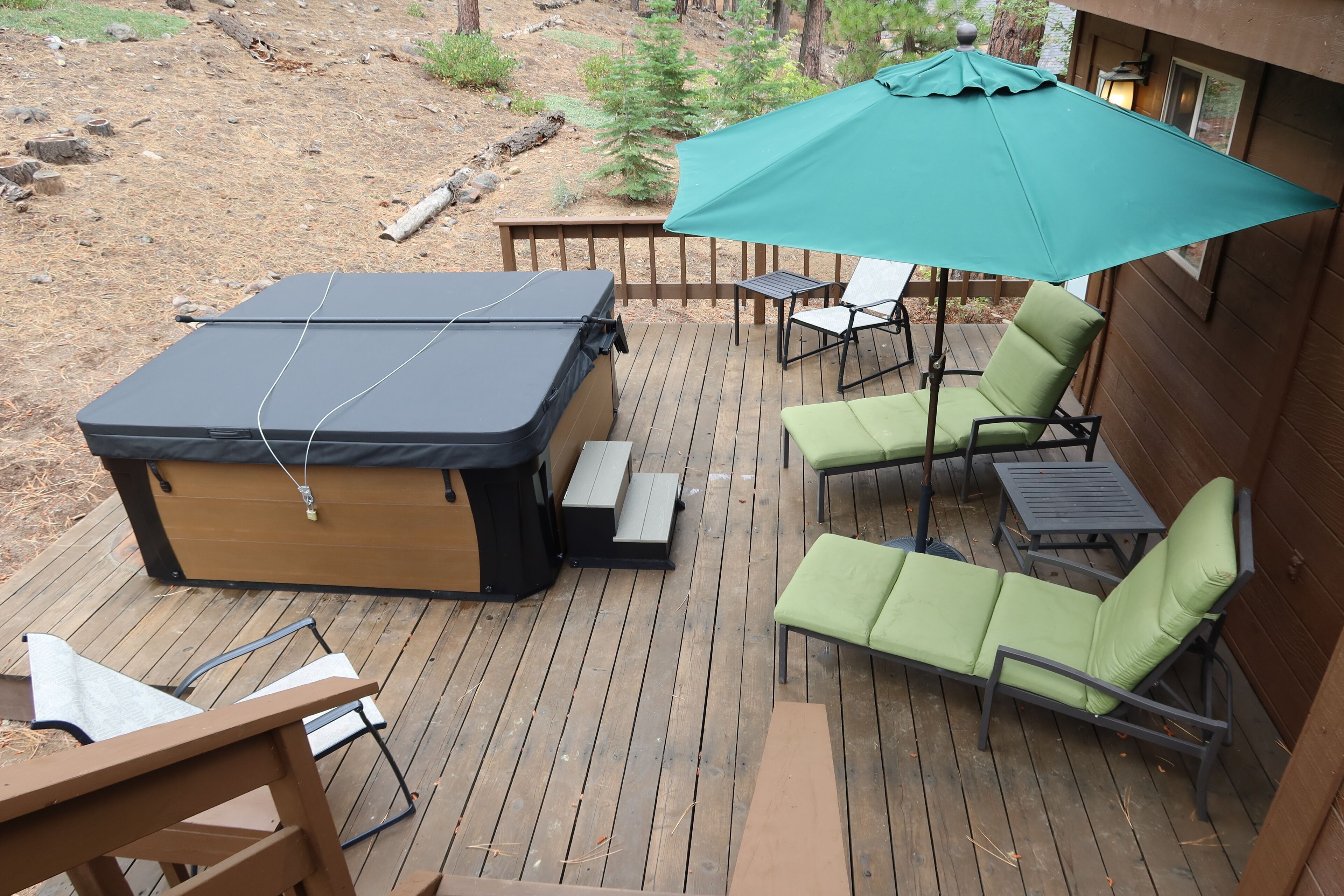 A deck with a hot tub, green lounge chairs, and a table under a large umbrella.