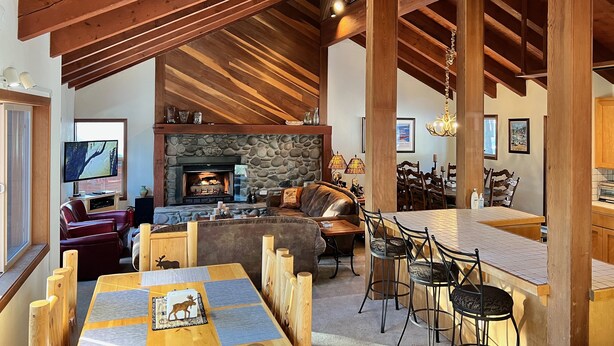 A spacious living area with a stone fireplace, wooden ceiling beams, and a cozy seating arrangement. - Serene Vistas at Northstar (Truckee)