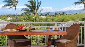 A wooden table with wicker chairs on a balcony overlooking a tropical landscape with palm trees and a view of the ocean.