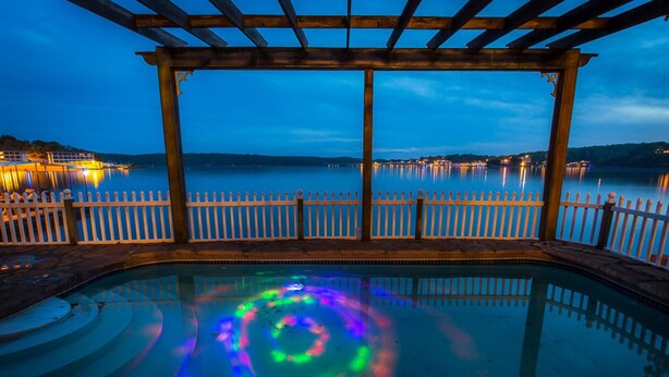 A pool with a wooden pergola and white fence, surrounded by calm water and distant city lights.