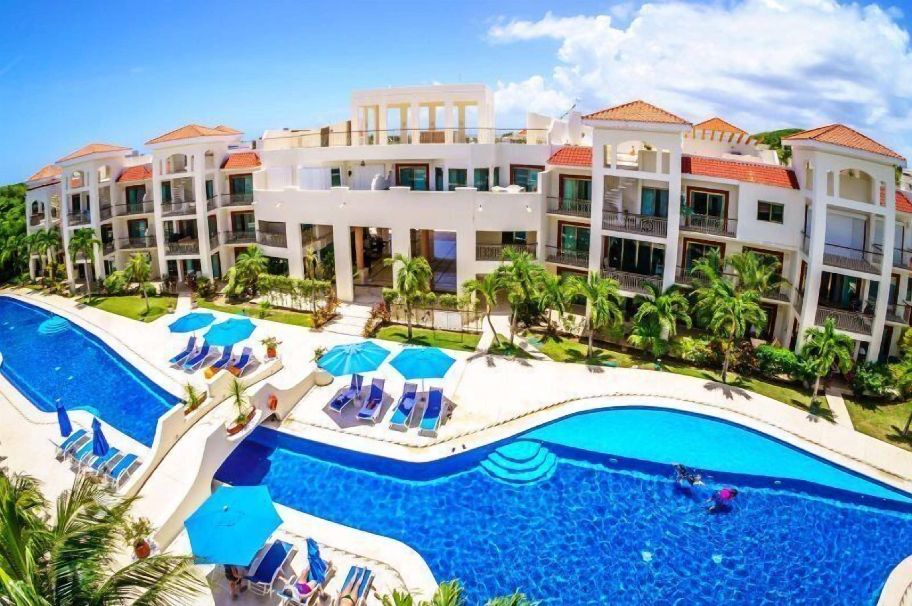 A resort with a large swimming pool, multiple balconies, and palm trees.