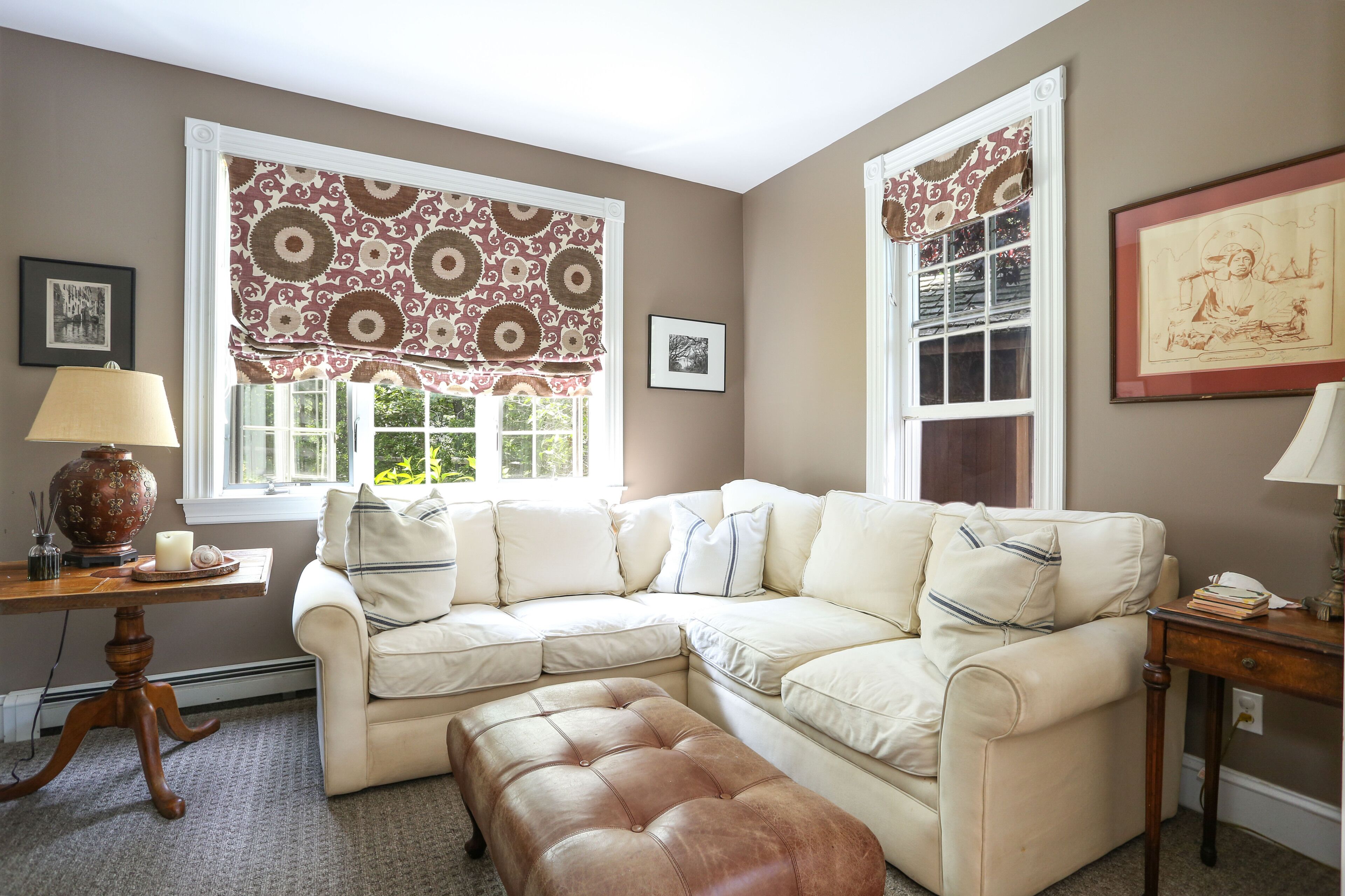 A living room with a beige sectional sofa, a brown leather ottoman, a wooden side table, and a window with patterned curtains.