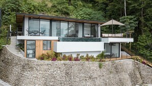Une maison moderne dotée d’un mur de soutènement en pierre, de grandes fenêtres en verre et d’une terrasse en bois.