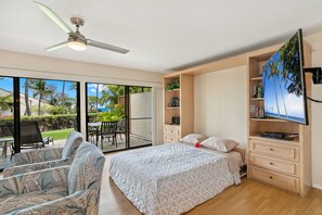 Une chambre à coucher comprenant un lit, un téléviseur, un ventilateur de plafond, une porte-fenêtre coulissante donnant sur un balcon et une commode en bois.