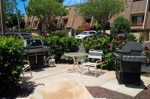 Relax in a serene outdoor space with a barbecue grill, round table, and comfortable bench amidst lush greenery and flowering plants.