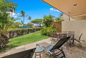 An outdoor patio with a table and chairs, a stone floor, and a view of a lush garden and palm trees.
