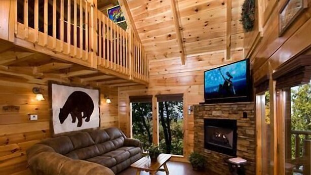 L’intérieur d’un chalet en bois est doté d’une cheminée en pierre, d’un canapé en cuir et d’un téléviseur fixé au mur.