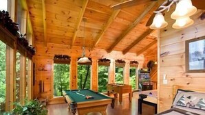 L’intérieur d’un chalet en bois comprend une table de billard, un lit recouvert d’une courtepointe et de grandes fenêtres donnant sur la forêt environnante.