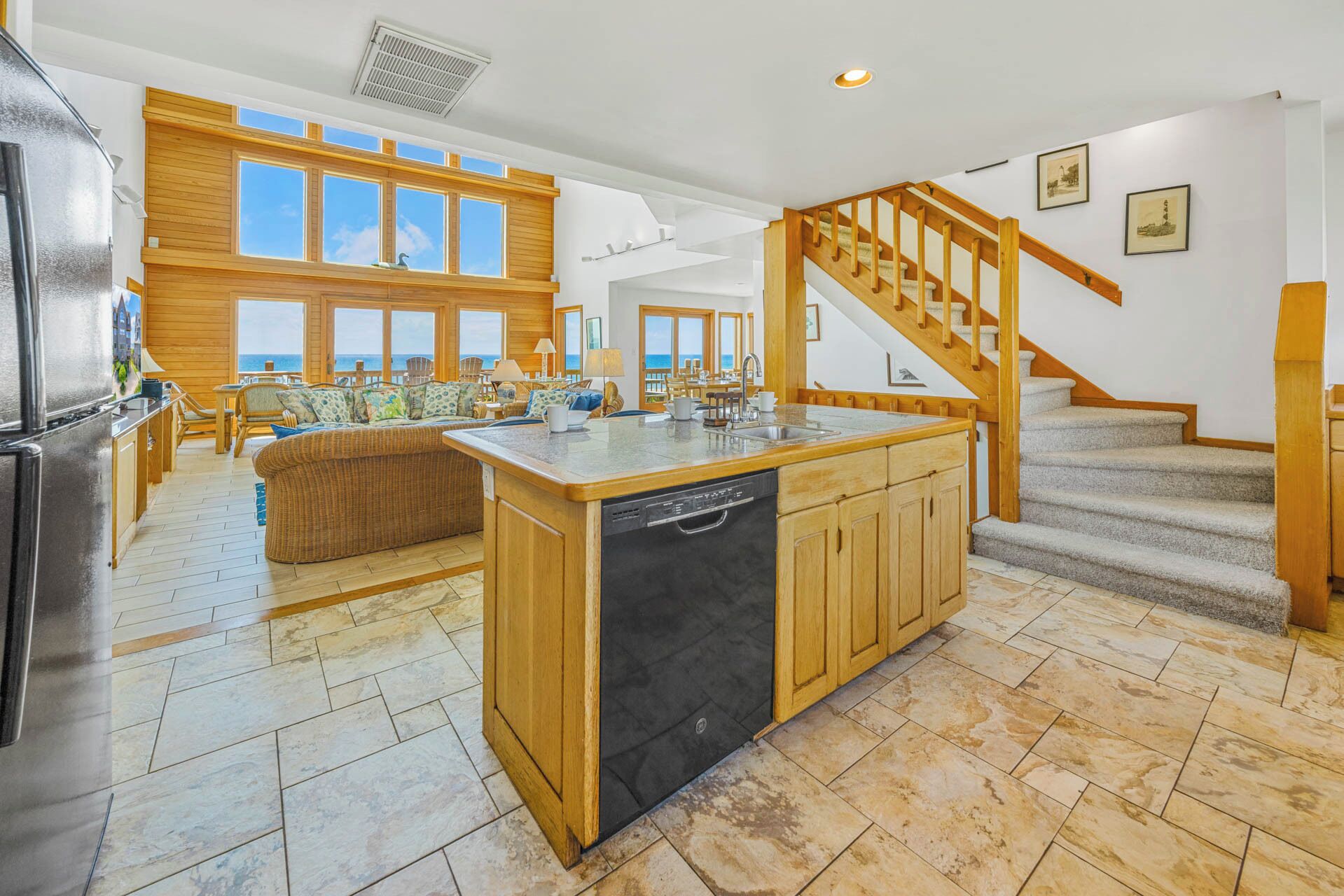 A spacious kitchen with a central island, wooden cabinetry, and a dishwasher. A wooden staircase leads to a balcony with a view of the ocean.