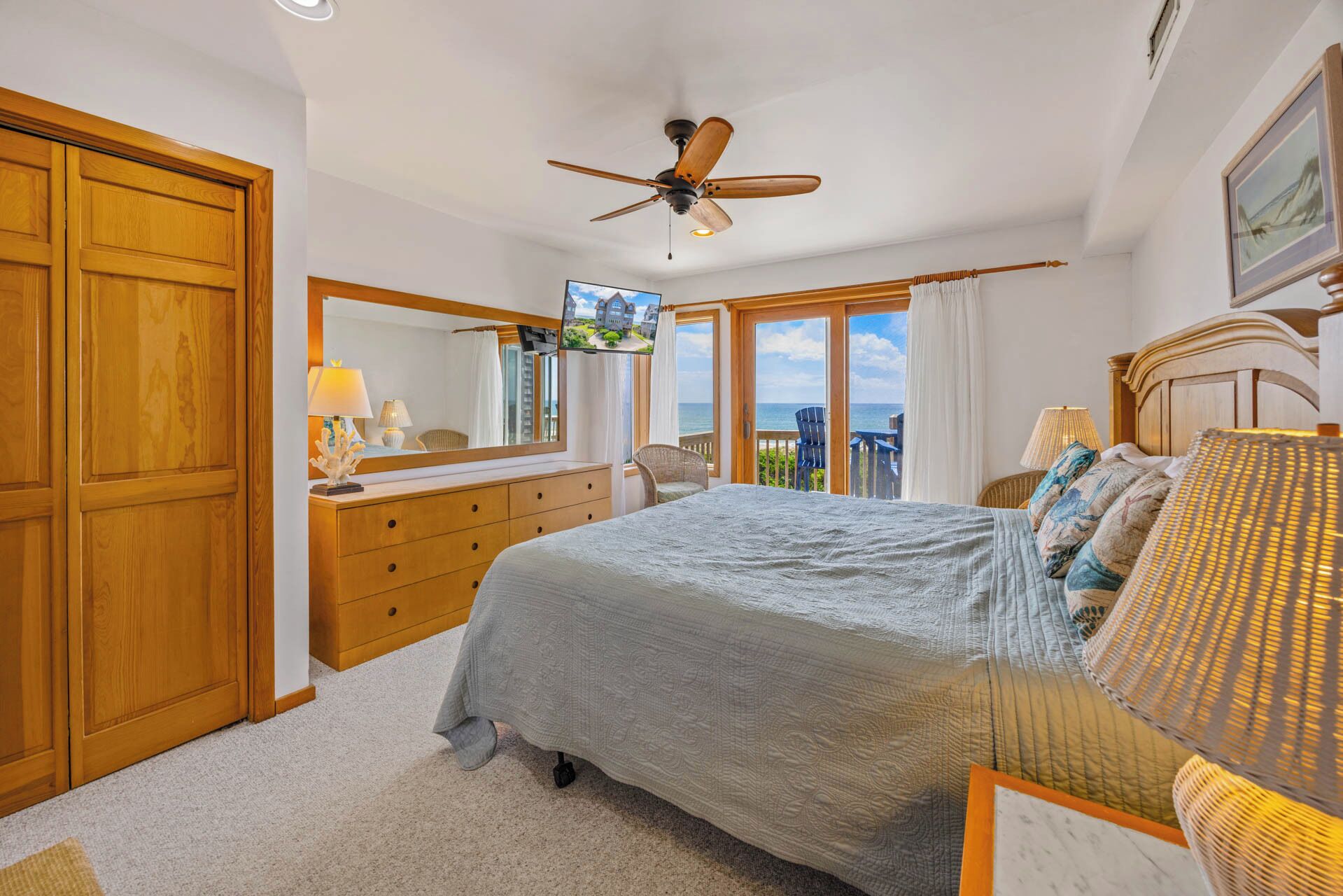 A bedroom with a large bed, a wooden wardrobe, a dresser, a ceiling fan, and a balcony with a view of the sea.