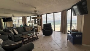 Un salón amplio con un ventanal grande con vistas a la playa, un ventilador de techo y un televisor de pantalla plana montado en la pared.