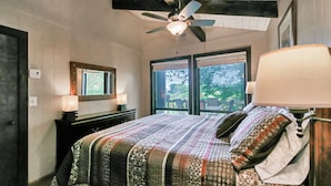 A bedroom with a bed, a ceiling fan, a window with a view of greenery, and a bedside lamp.