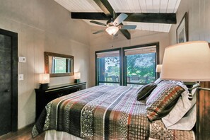 A bedroom with a bed, a ceiling fan, a window with a view of greenery, and a bedside lamp.