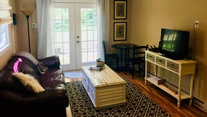A living room with a sofa, a coffee table, a TV on a stand, a dining table with chairs, and a door with curtains. - Relaxing Cottage close to everything! Perfect for 2!Wi-Fi, Keurig,Outside space! (Greensboro)