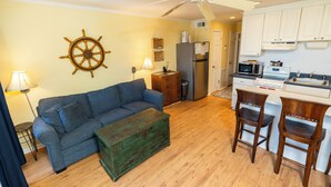 Un espace de vie compact comprenant un canapé, une table basse, une commode en bois, un ventilateur de plafond et un coin cuisine équipé d’un réfrigérateur, d’un four à micro-ondes et de tabourets de bar.