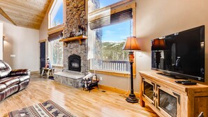 A spacious living room with a stone fireplace, wooden flooring, a leather sofa, a large flat-screen TV, and a view of the outdoors through large windows.