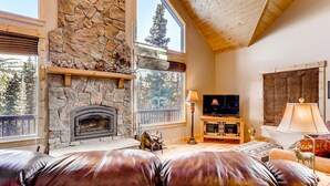 A cozy living room with a stone fireplace, leather seating, a television, and a large window offering a view of trees.