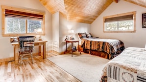 A cozy bedroom with a wooden ceiling, a bed with a patterned bedspread, a desk with a chair, and a window with a view of trees.