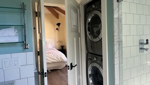 Un cuarto de lavado con dos lavadoras, una zona de ducha y una cama visible a través de un umbral abierto.