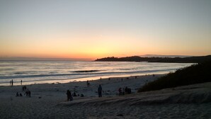 Una playa con gente caminando y sentada, un atardecer de fondo y una costa distante.