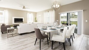 Un salon moderne avec un canapé blanc, une table basse en verre et un téléviseur à écran plat. On y trouve également un coin repas avec une table blanche dressée pour six personnes, entourée de chaises blanches.