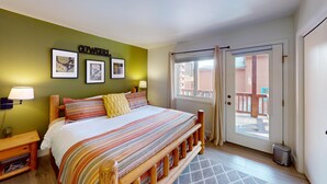 Un dormitorio con cama, mesita de noche, lámpara de pared y puerta que da a un balcón exterior.