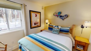 Un dormitorio con cama, mesita de noche, lámpara, un cuadro enmarco y ventana con cortinas.