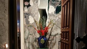 Ein Eingangsbereich mit einer dekorativen Vase auf einem Sockel, einer Fußmatte und einer Tür vor einer strukturierten Wand.