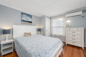 Un dormitorio con una cama, una mesita de noche con lámpara, un aparador, un cuadro en la pared y un ventanal con cortinas.