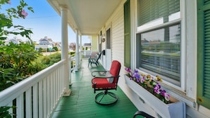 Une véranda avec un plancher vert, une balustrade blanche et une chaise rouge. On y trouve des fenêtres avec des volets verts et une jardinière ornée de fleurs violettes et jaunes.