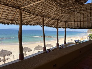 A beachside pavilion with a thatched roof overlooking a sandy beach and the sea.