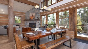 Interior rústico de una cabaña con chimenea de piedra, mesa de comedor de madera, bancos y ventanas grandes que ofrecen vistas al bosque.