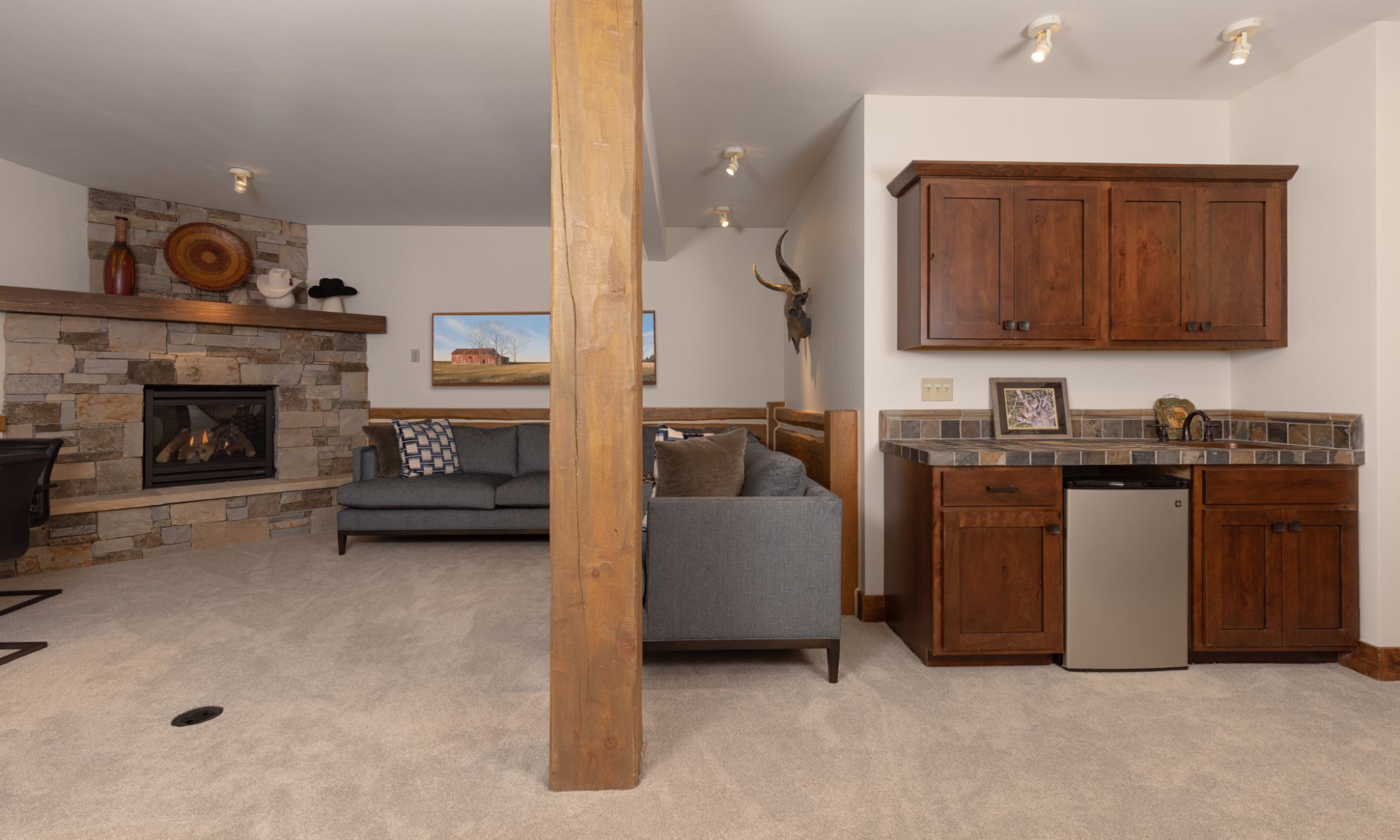 A living room with a stone fireplace, a gray sofa, and a kitchen with wooden cabinets and a dishwasher.