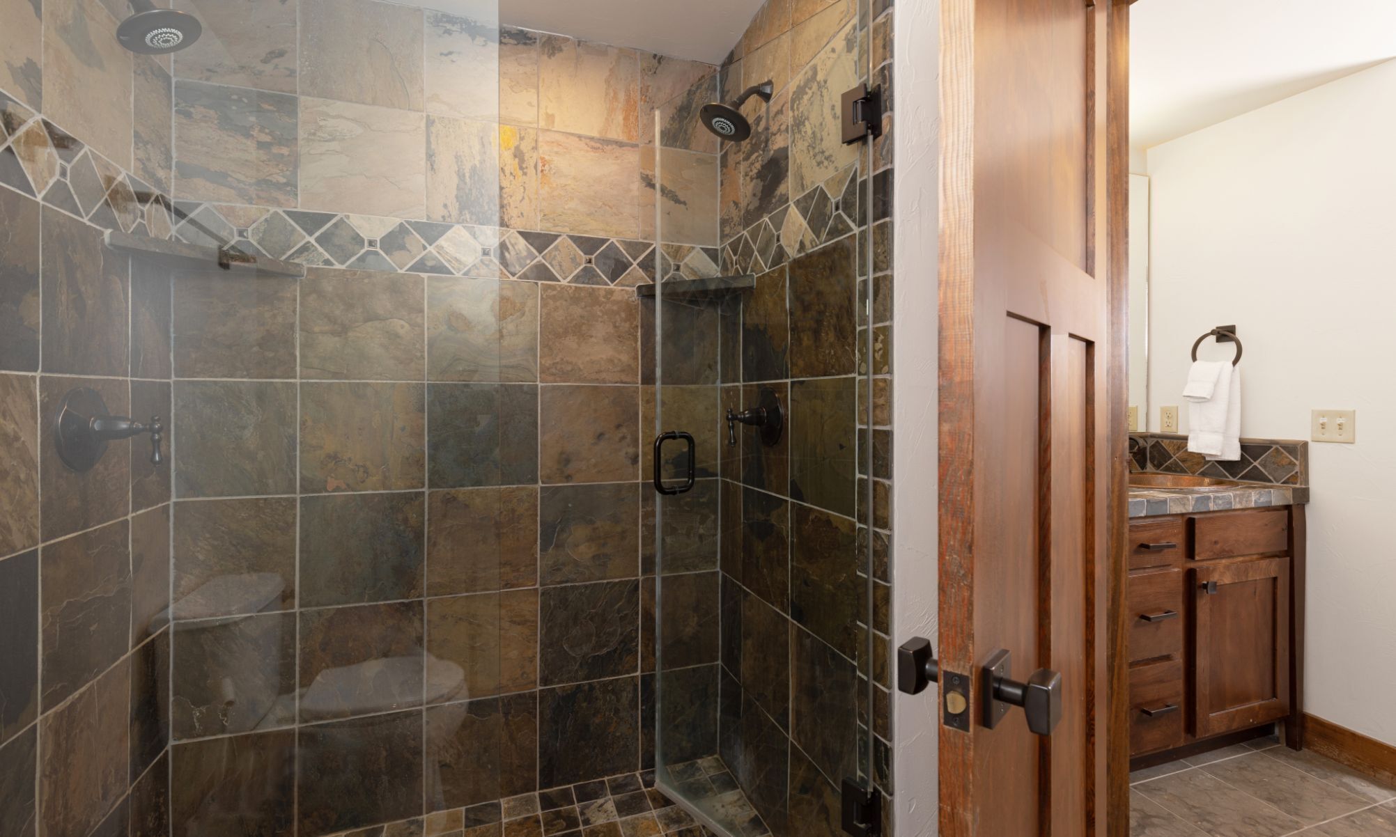 A shower with a glass door, stone tiles, and a wooden door leading to a bathroom with a sink and towel rack.