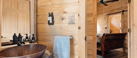 Een houten cabin met een grote houten spoelbak, een zeepdispenser in de vorm van een beer, een handdoek, een bed en een plafondventilator.