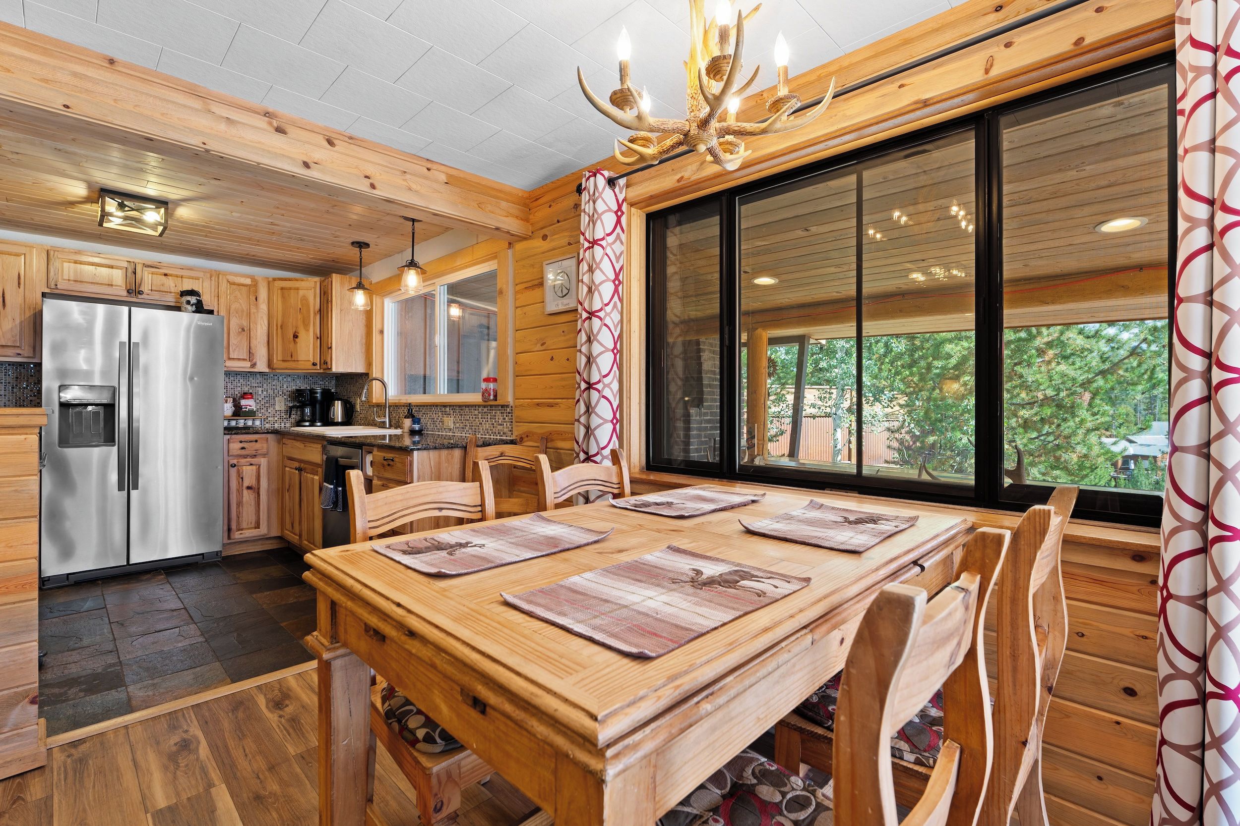 A wooden cabin interior with a dining table, chairs, a refrigerator, and a large window.
