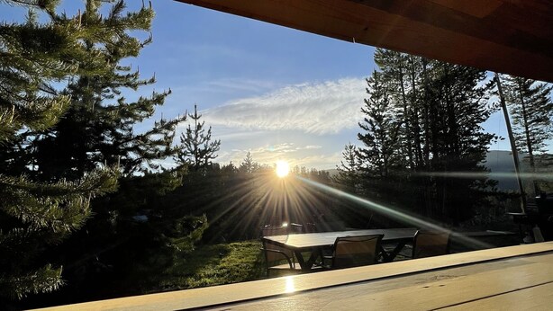 Un pontile in legno con un design circolare, affacciato su una zona boschiva con cielo sereno e il sole che tramonta in lontananza.